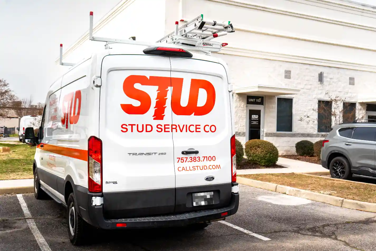 A white van with “STUD STUD SERVICE CO” and contact information printed in large orange letters parked outside a commercial building.
