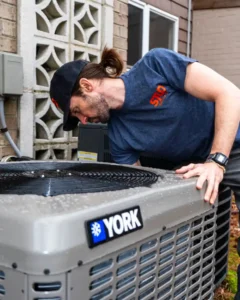 A man in a blue shirt and cap inspects a YORK outdoor air conditioning unit next to a building.