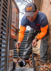 A technician in a STUD Services Co. uniform works on an outdoor HVAC unit, adjusting a valve with tools next to a brick wall.