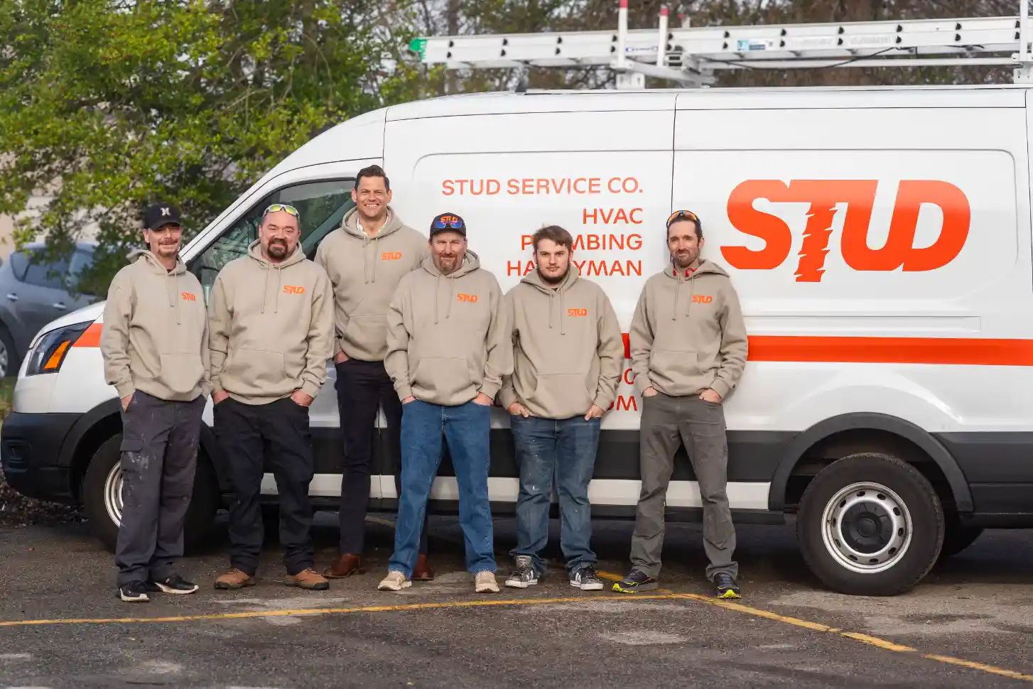 Six men wearing matching hoodies stand in front of a white service van with "STUD Service Co." signage, parked outdoors near some trees.