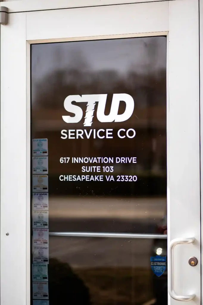 Glass door with “STUD SERVICE CO” and address, 617 Innovation Drive, Suite 103, Chesapeake VA 23320, in white lettering; several stickers are visible on the left side of the door.